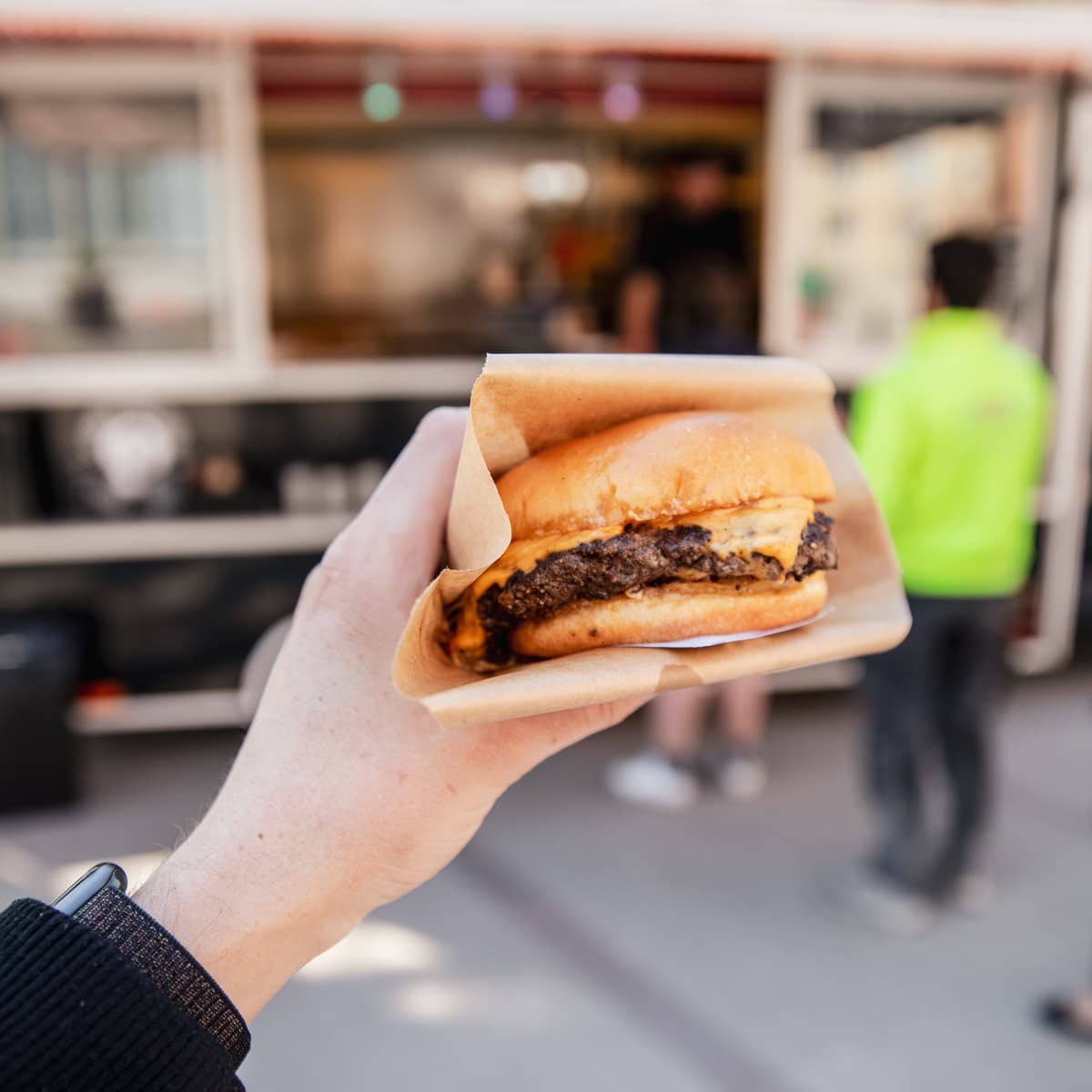 En person som håller en hamburgare framför en foodtruck.